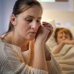 Young mother sitting next to bed of her sleeping son suffering from depression and bad thoughts. Concept of lonely mother, family problems and maternal depression