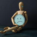 Wooden man with sad emoji in hands on dark blue background