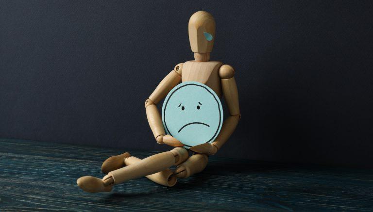 Wooden man with sad emoji in hands on dark blue background