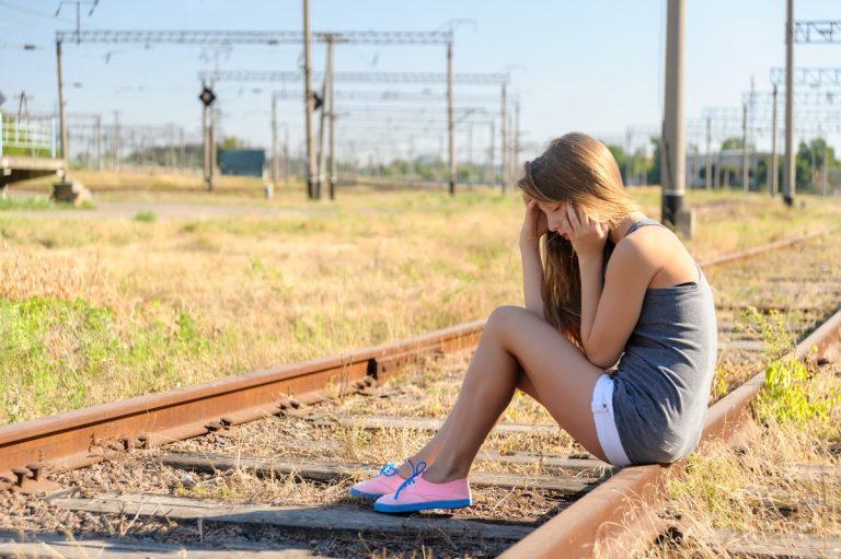 Upset teenager girl sitting on rail track in countryside
